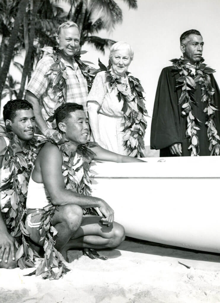 Black and white photo of people standing near a canoe, wearing maile lei