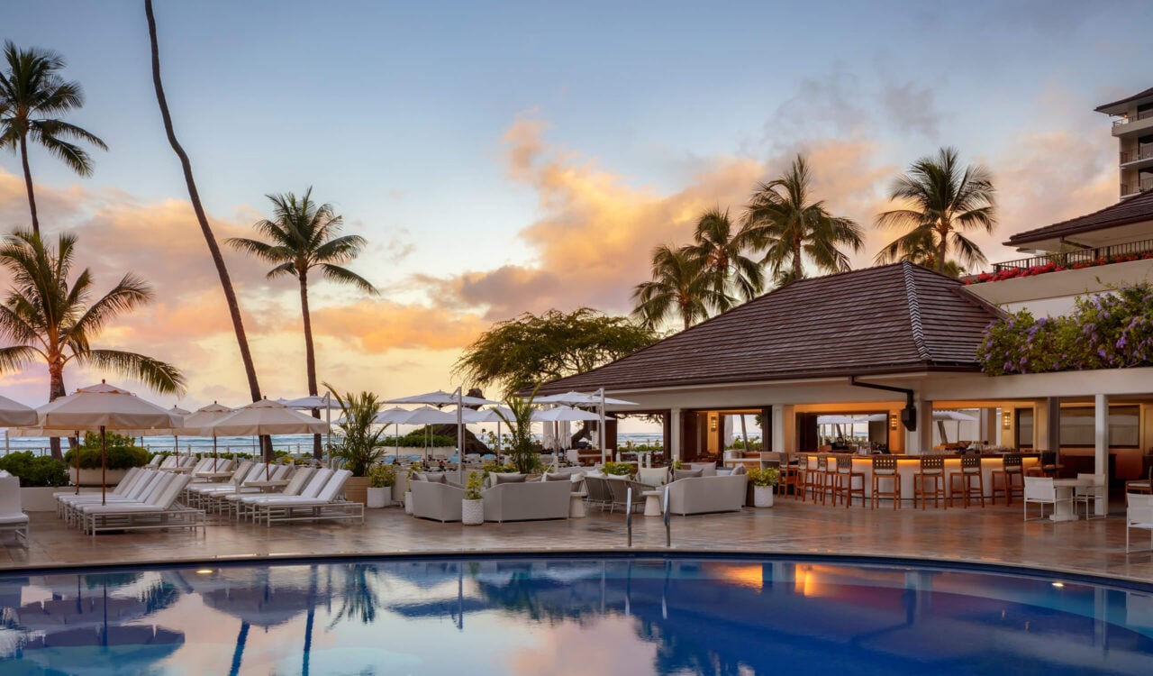 A luxurious resort pool area at sunset, featuring white lounge chairs, umbrellas, a poolside bar with seating, palm trees, and views of the ocean in the background.