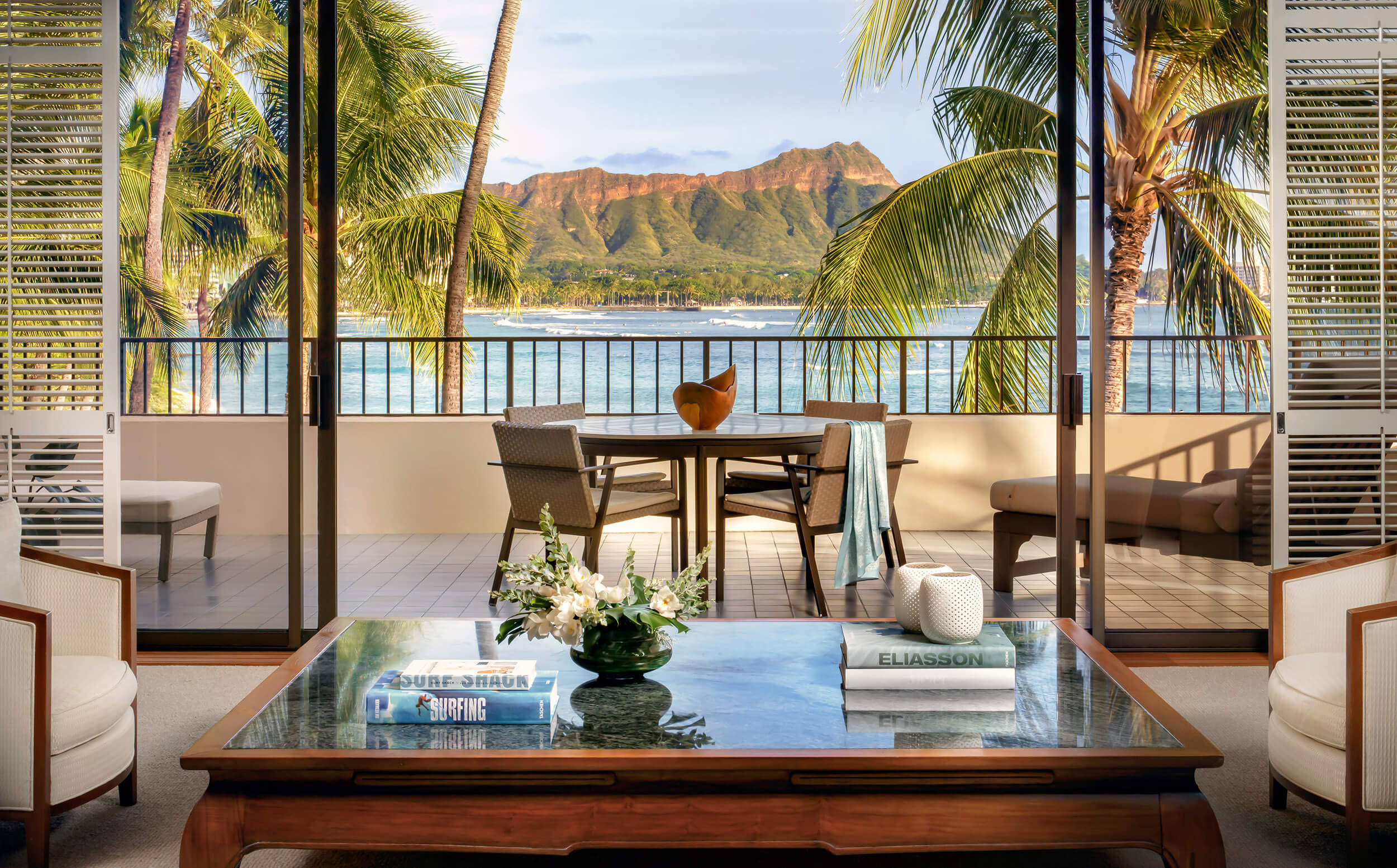 A living room with a glass-topped coffee table, two white chairs, and a balcony with a view of the ocean and palm trees.